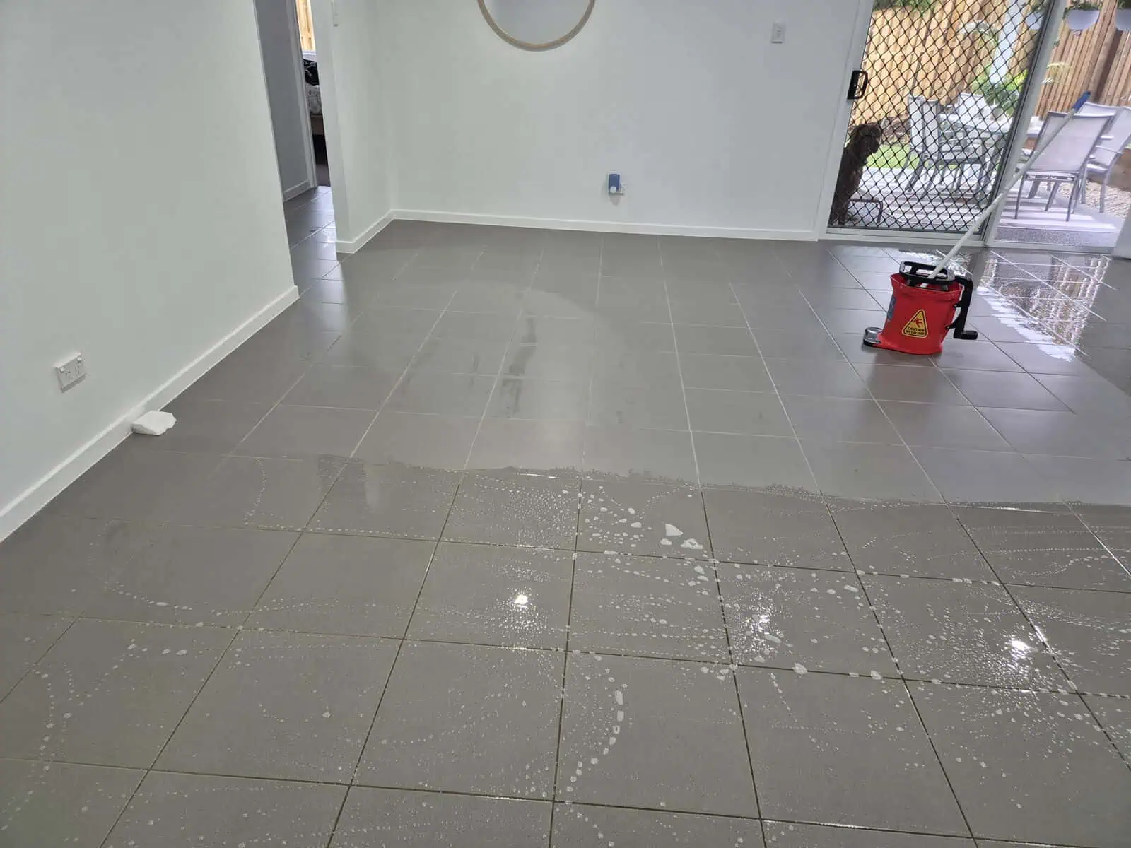 Grey tiles halfway through being mopped with red mop bucket sat on floor